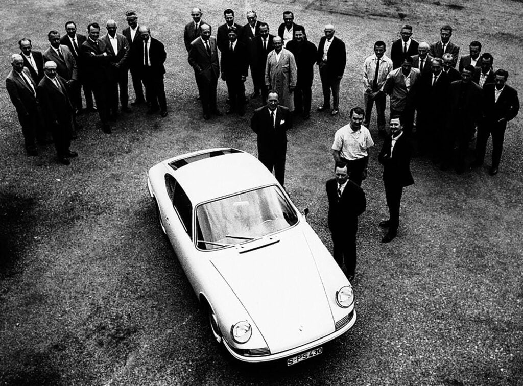 black and white photo of groups of people standing next to a Porsche car