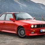 Red BMW E30 M3 parked on a cliffside with a mountain behind it