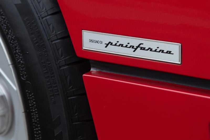 silver badge with black writing on it that says Disegno Di Pininfarino on a red car, tire and wheel to the left of it