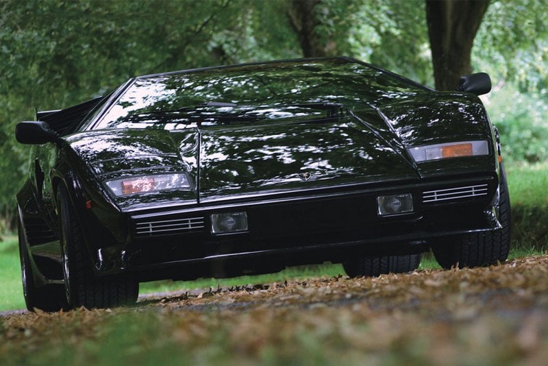 Black Countach driving in a forest with leaves on the ground and trees behind it