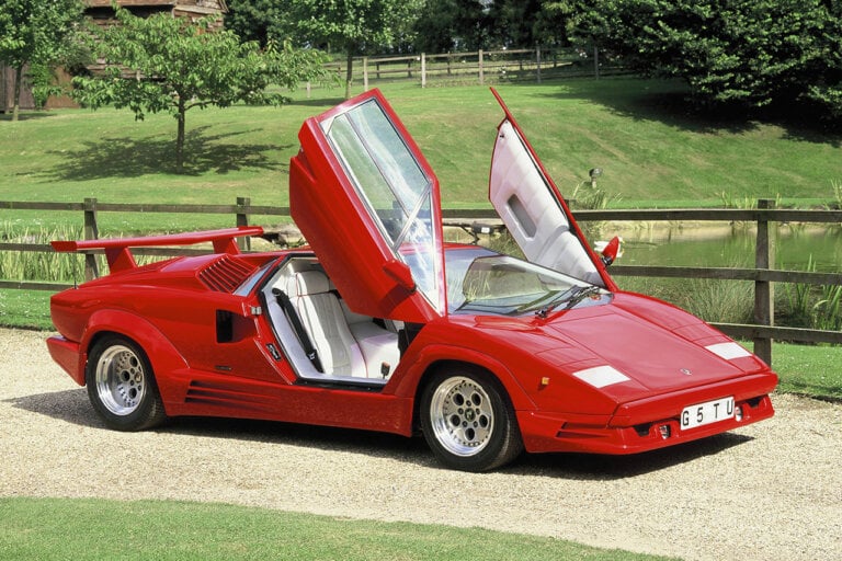 red Lamborghini Countach with scissor doors open and wood fence running behind it. Large grassy field behind both of those objects