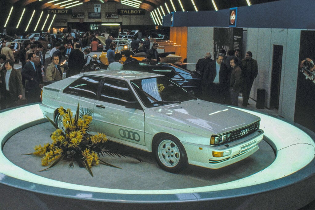 White Audi Quattro sitting on a platform with a white ring around it and many people surrounding the car