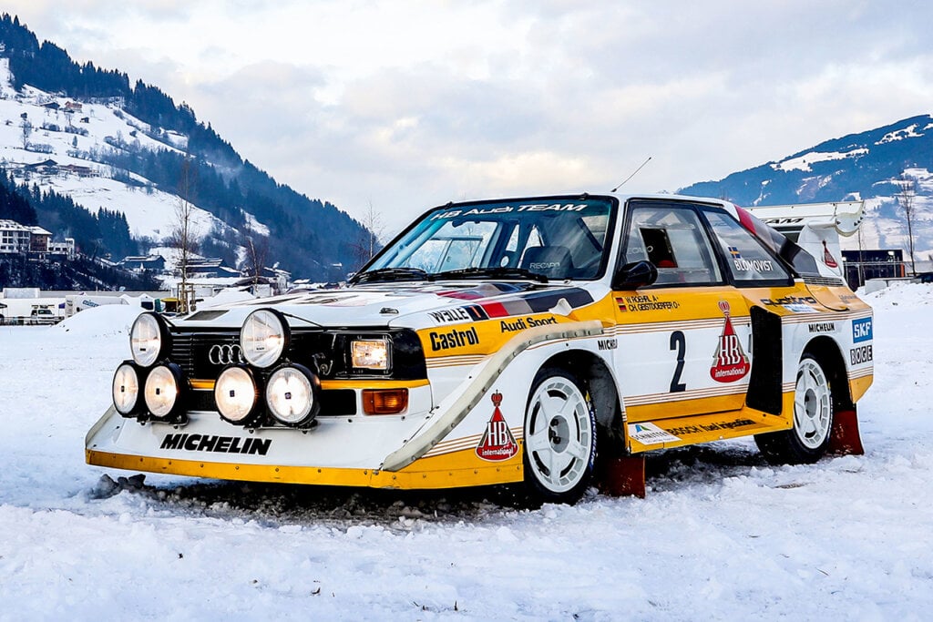 White Audi Sport Quattro S1 E2 with yellow stripes parked with lights on in the snow with mountains behind it