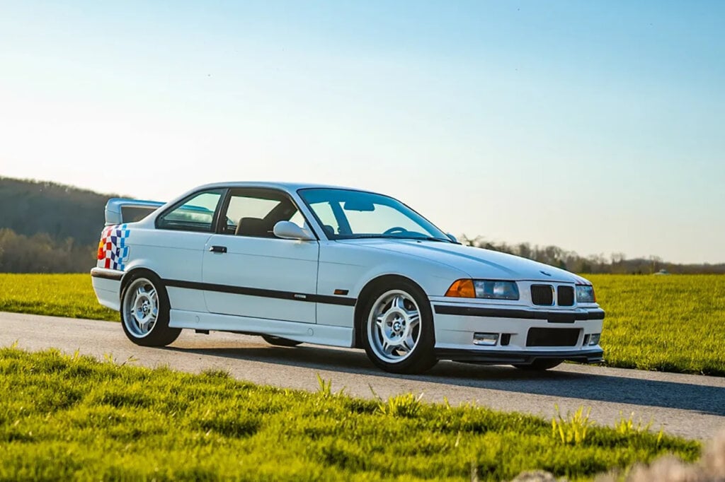 white Lightweight BMW e36 m3 with rainbow checkers on back of car parked on strip of asphalt next to grass and clear sky in background