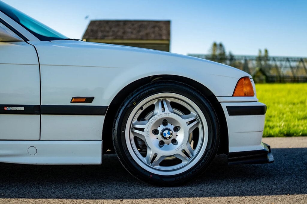 5 spoke wheel on a white BMW E36 M3 lightweight variant. Grass and large building behind the car.