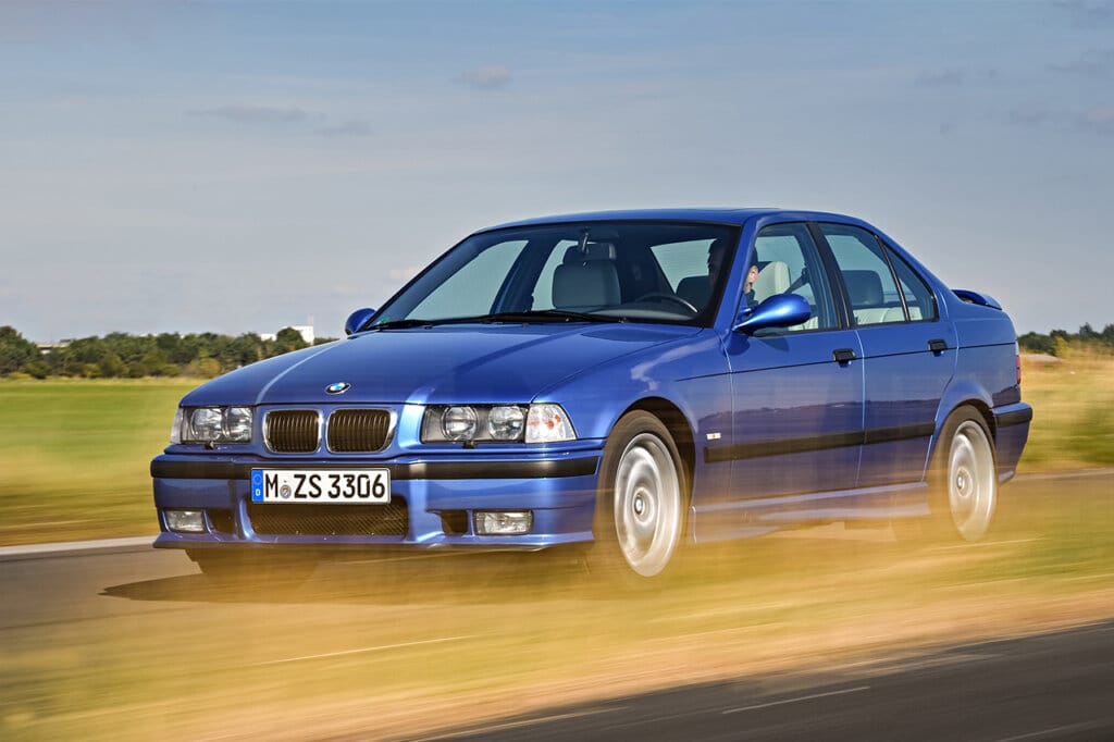 Blue m3 e36 on road near a grass field. Grass infront of the car is passing it really fast