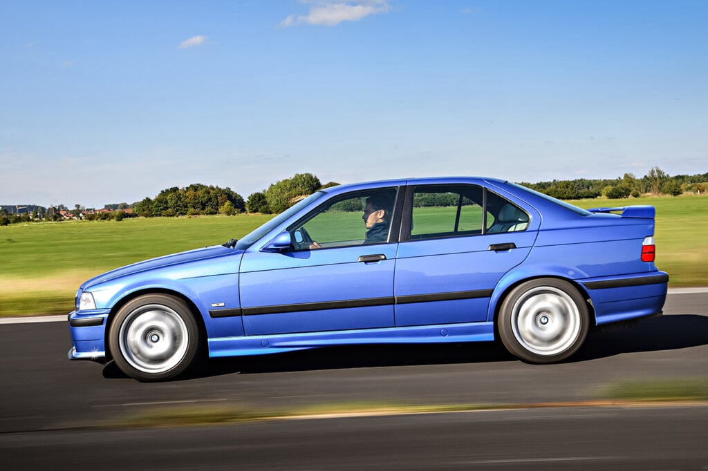 Blue BMW driving on a long strip of road with grass to the right side of the car and clear blue sky