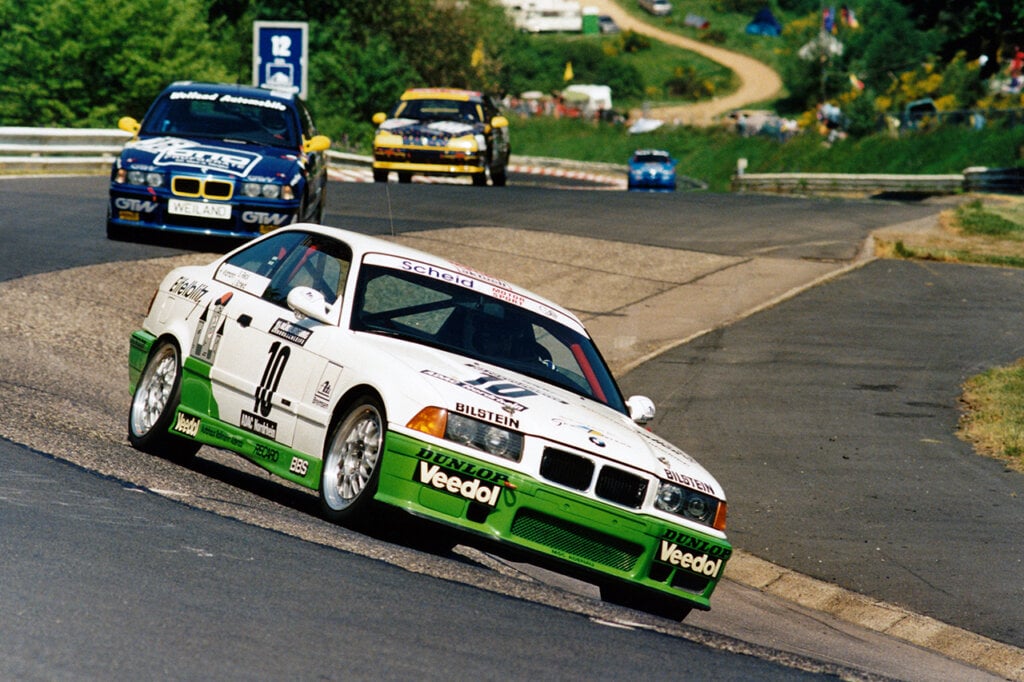 white and green BMW e36 M3 with race livery turning through a turn with other cars following closely behind