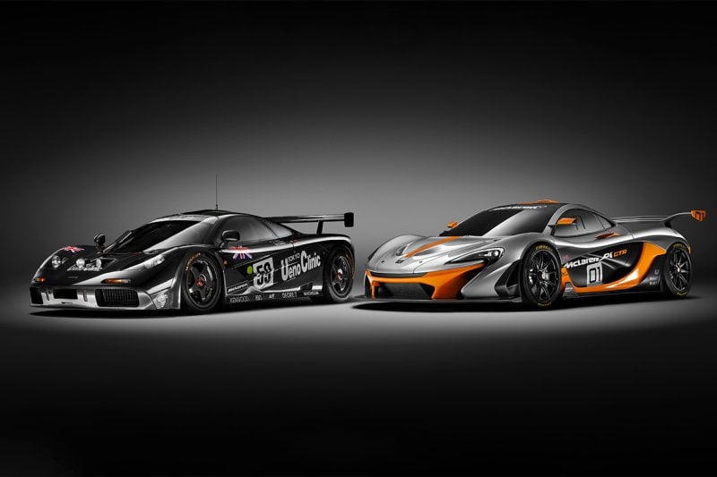 Black McLaren F1 with Ueno Clinic race livery next to an orange McLaren P1. Both parked with a spotlight shining on the car and black background