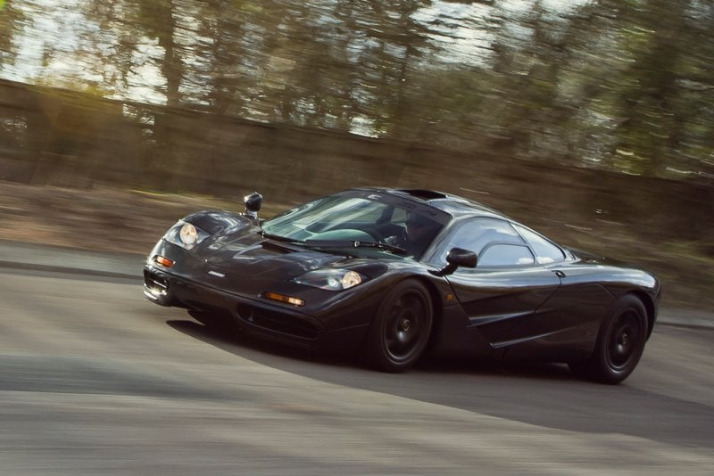 Black McLaren F1 driving fast on the road. Sun shining through trees in background
