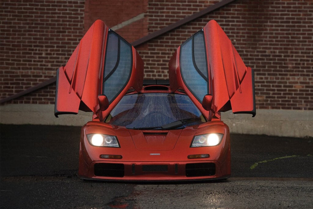 Red McLaren F1 parked in the rain, doors open and headlights on in front of brick building