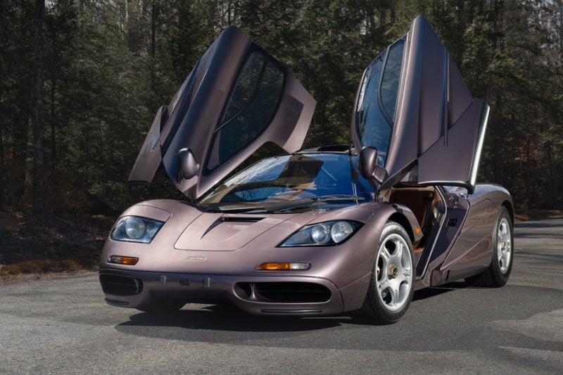 Purple McLaren F1 with doors open parked on the side of a road with trees in background