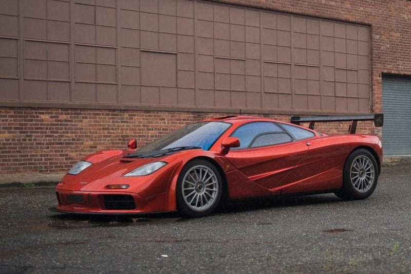 Red McLaren F1 with raindrops on it parked on wet ground in front of brick red building