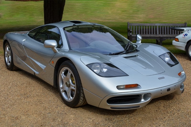 Silver McLaren F1 parked on rocks and green grass behind it. Parked next to a white car