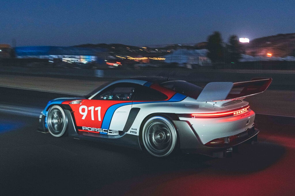 Porsche 911 GT3 R Rennsport driving on a road passing buildings in the background at night