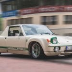 White with green trim Porsche 914 driving on a road passing a building