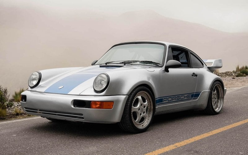 silver Porsche 911 Carrera RS 3.8 with blue racing stripes next to a cliff
