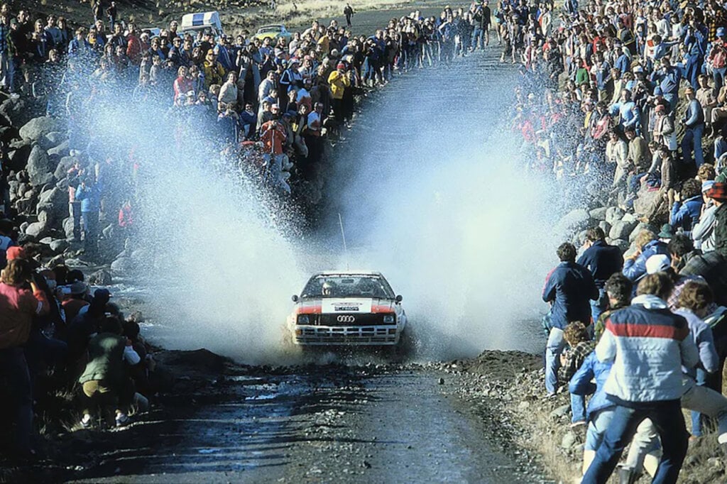 Audi Rally Race car driving through a stream of water and spraying huge mist of water onto spectators surrounding the course