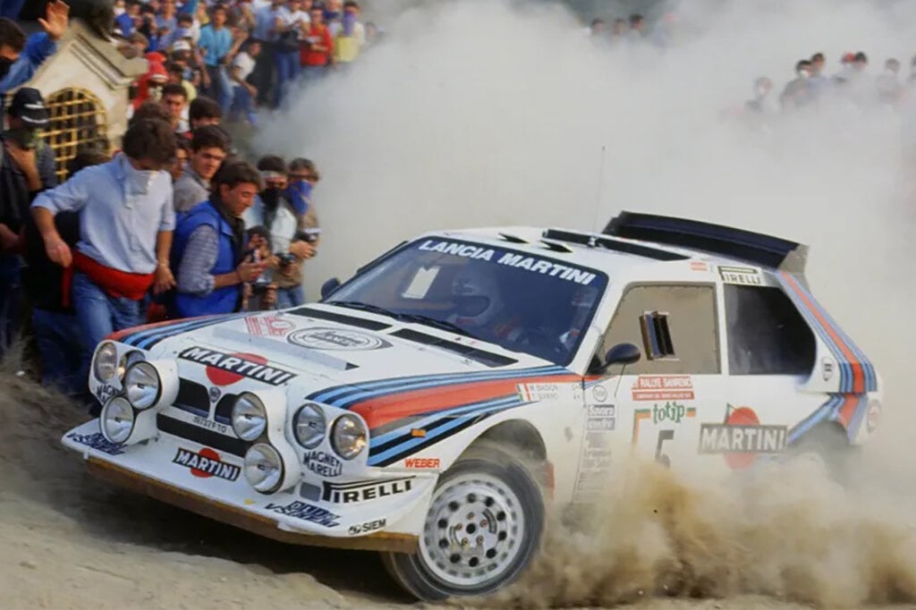 White Lancia Delta S4 turning hard in the dirt amongst a crowd of people