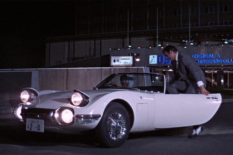 Man getting into a white Toyota 2000GT with a woman in the passenger seat