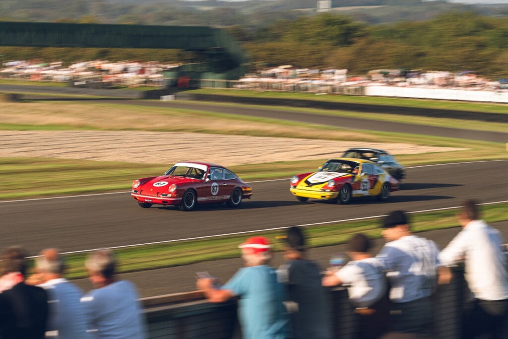 Porsche 911s one red and the other yellow, racing through a turn infront of spectators