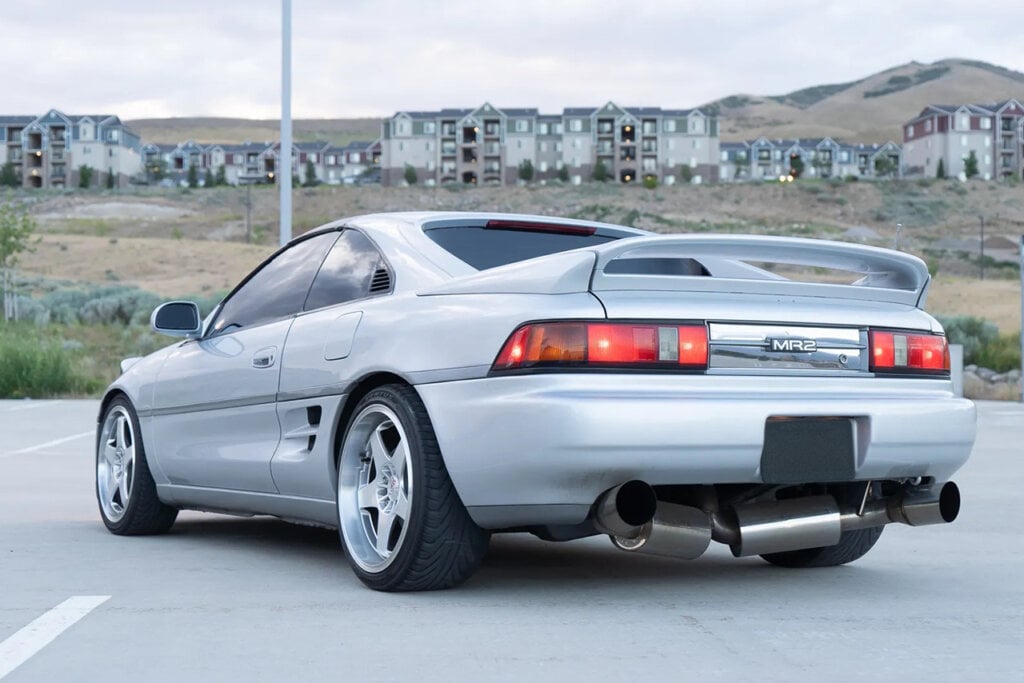 Rear side of a Silver Toyota MR2 with houses in the background