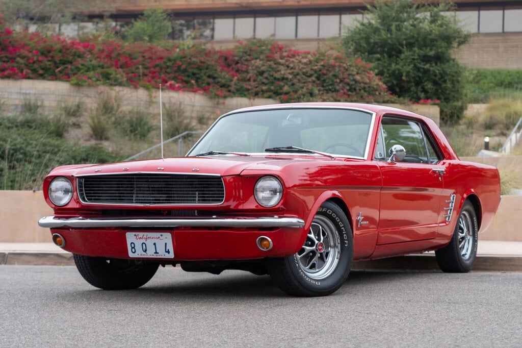 Red 1964 Ford Mustang parked infront of a curb and a building is in the background