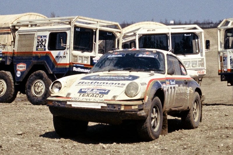 Dakar Porsche rally car parked in front of busses and trucks with the same paint job