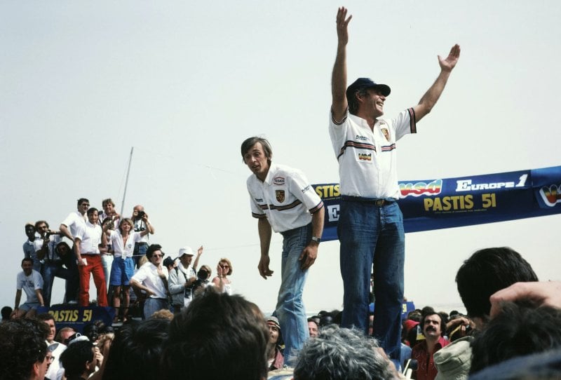 Two men standing on a car in the middle of a crowd of people, one with his arms raised to the sky
