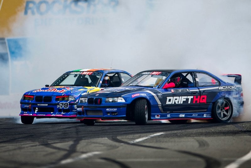 Two blue BMW E36 m3's drifting through a turn with a huge cloud of smoke behind them