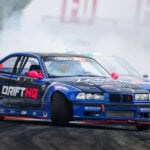 Two blue e36 drift cars close to one another on a road with tire marks and smoke in background