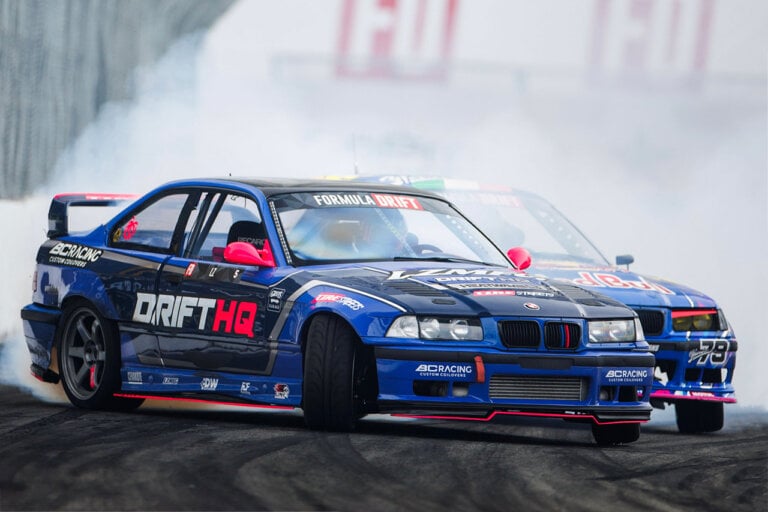 Two blue e36 drift cars close to one another on a road with tire marks and smoke in background