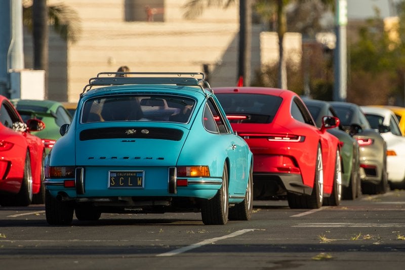Blue and red Porsches near other cars behind one another in a parking lot