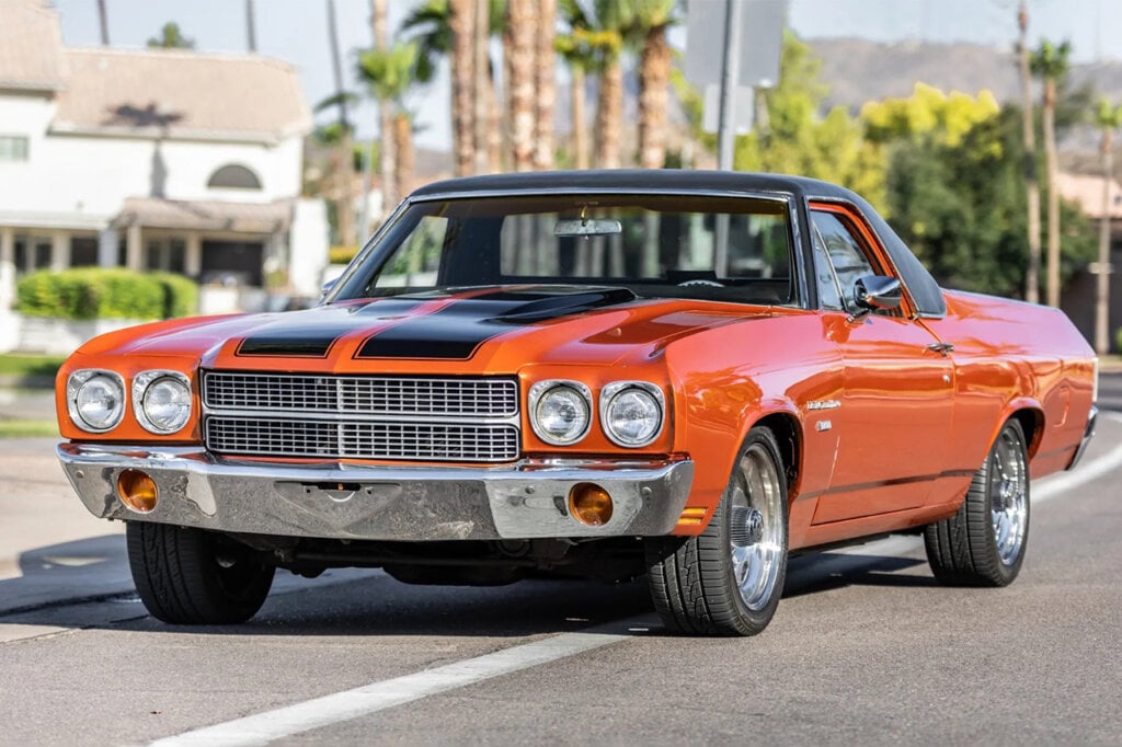 Orange and black Chevy El Camino parked on the side of a road with palm trees behind it