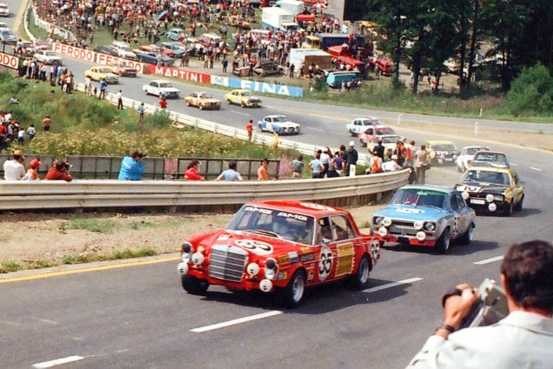 Color image of several race cars behind a red Mercedes 300SEL with spectators in background