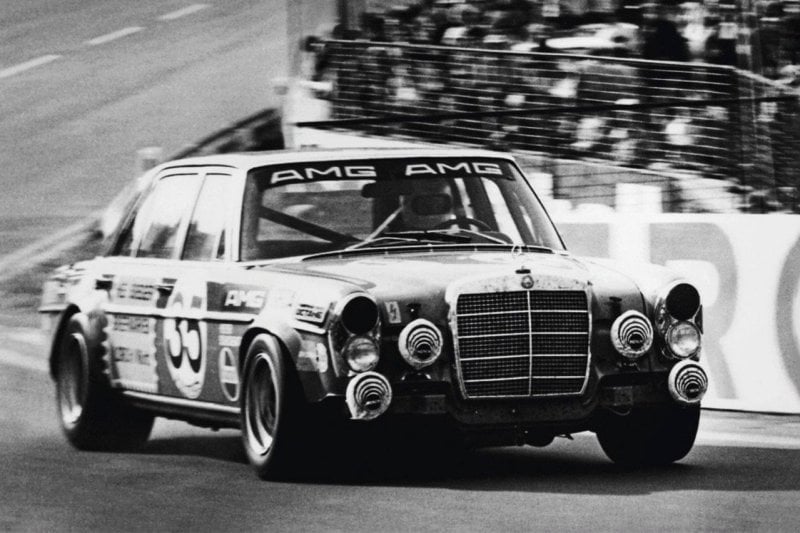 black and white photo of Red pig AMG 300SEL driving on a racetrack with specatators behind it