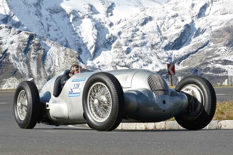 Classic silver race car with man driving it through snow covered mountain pass