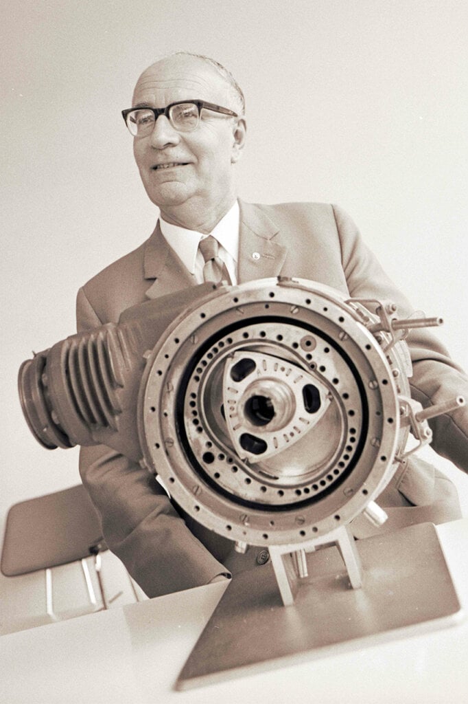 grey photo of man in a suit with glasses standing behind an engine part