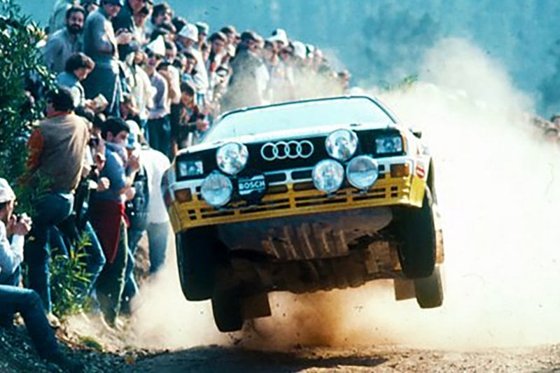 White and yellow Audi flying through the air leaving a dust cloud and passing spectators