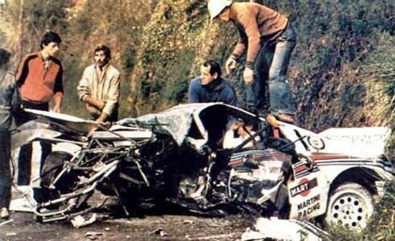 a white Group B rally car crumpled and broken with a man standing on top of it