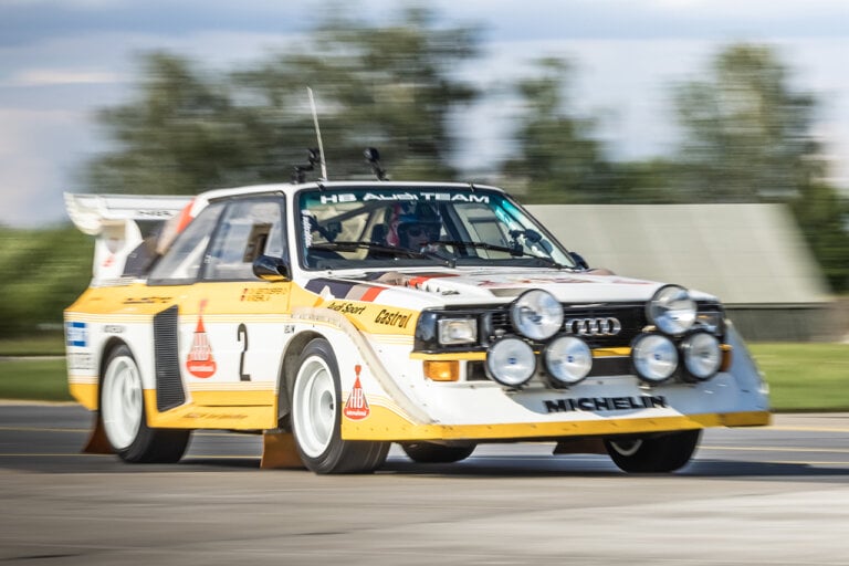 Yellow and white Audi Quattro with a man in a race suit driving past trees