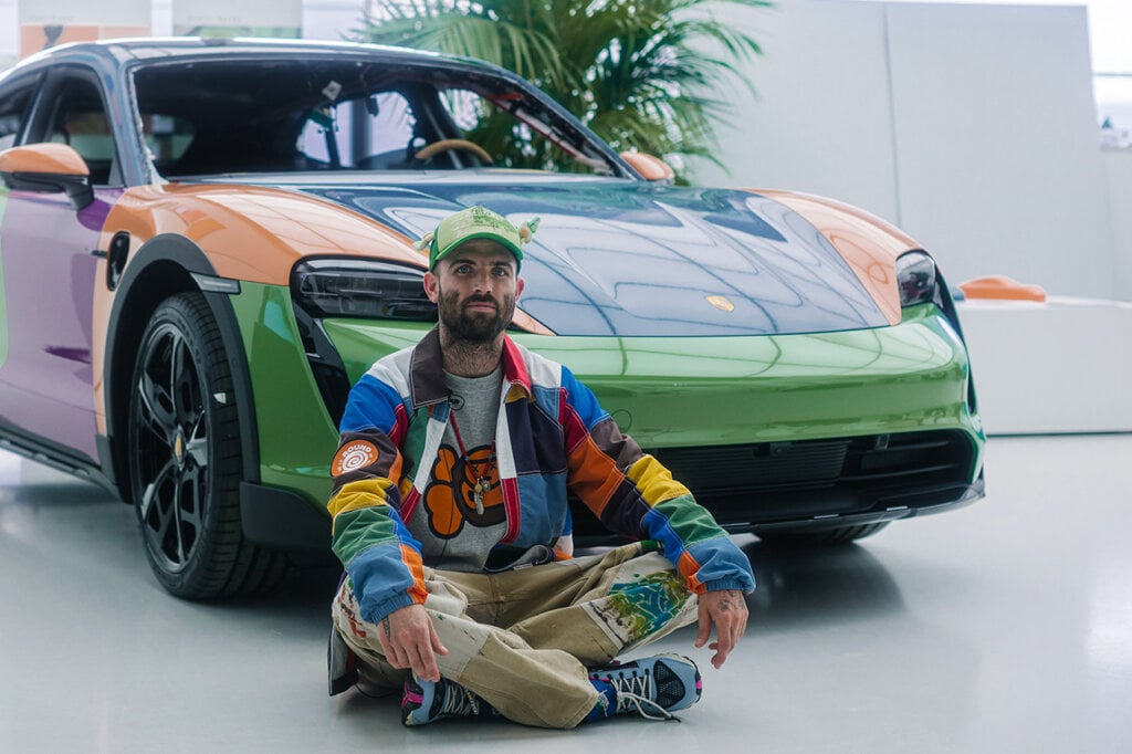 Man in multicolored jacket with a hat on sitting cross legged in front of a multi colored Porsche