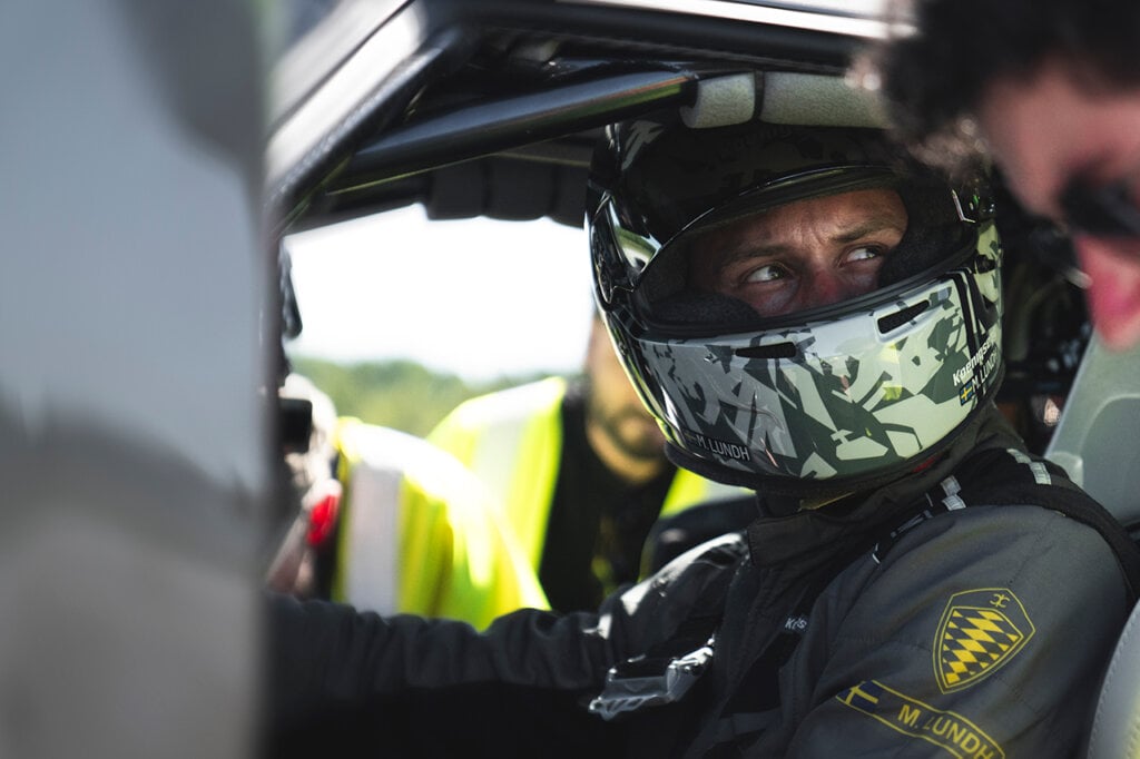 man with a racing helmet and race suit sitting in a car surrounded by people