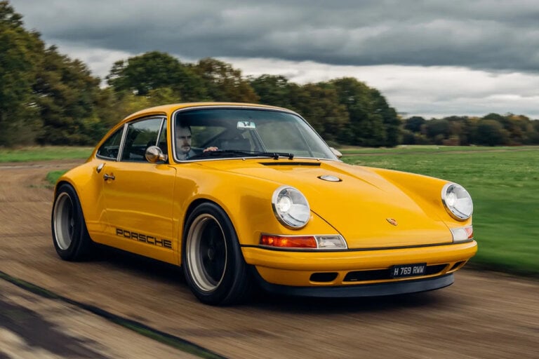 Yellow Singer Porsche UK1 with a man behind the wheel driving on a dirt road. With grey cloudy sky and green grass in the background.